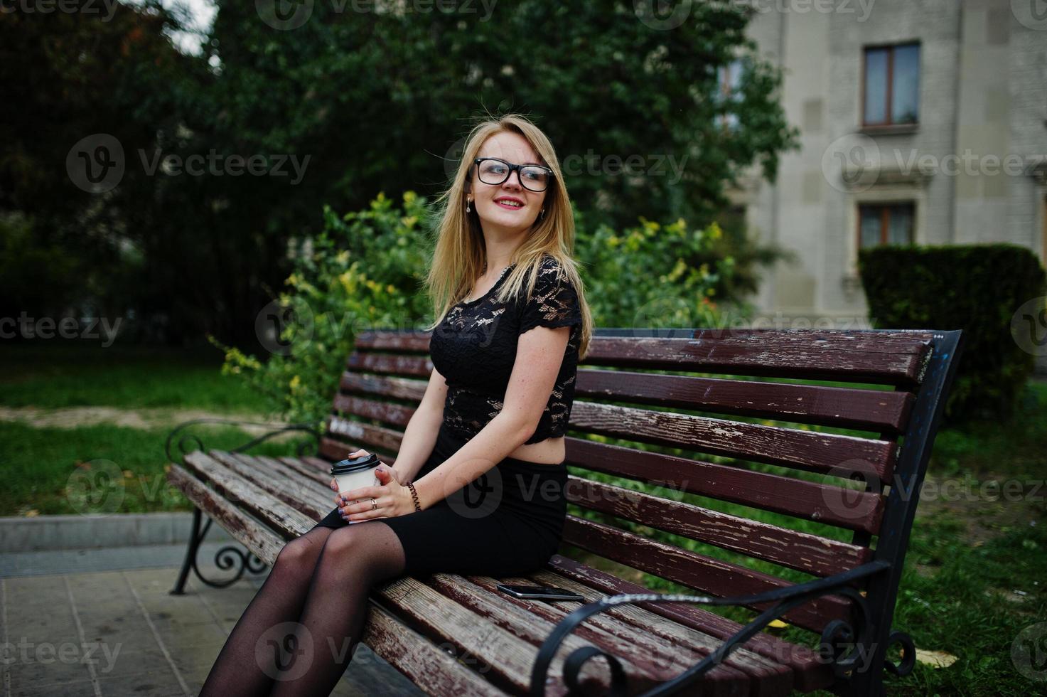 elegant blond meisje draagt zwarte poseren in de straten van de stad, zittend op een bankje met een kopje koffie bij de hand. foto