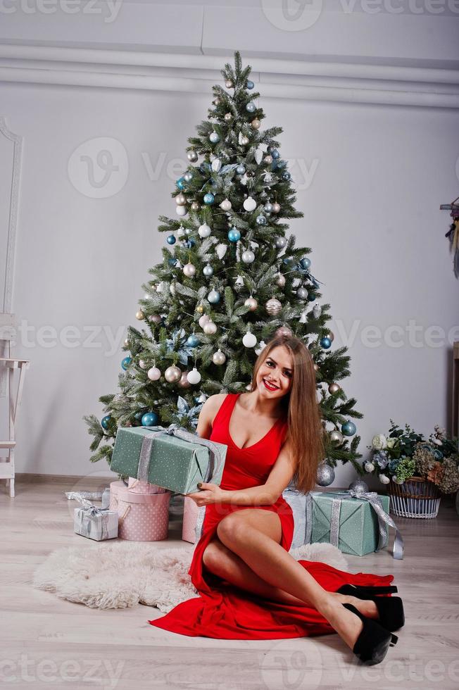 modieuze prachtige vrouw op rode lange avondjurk poseerde in studio tegen nieuwjaarsboom met geschenken. kerstvakantie thema. foto