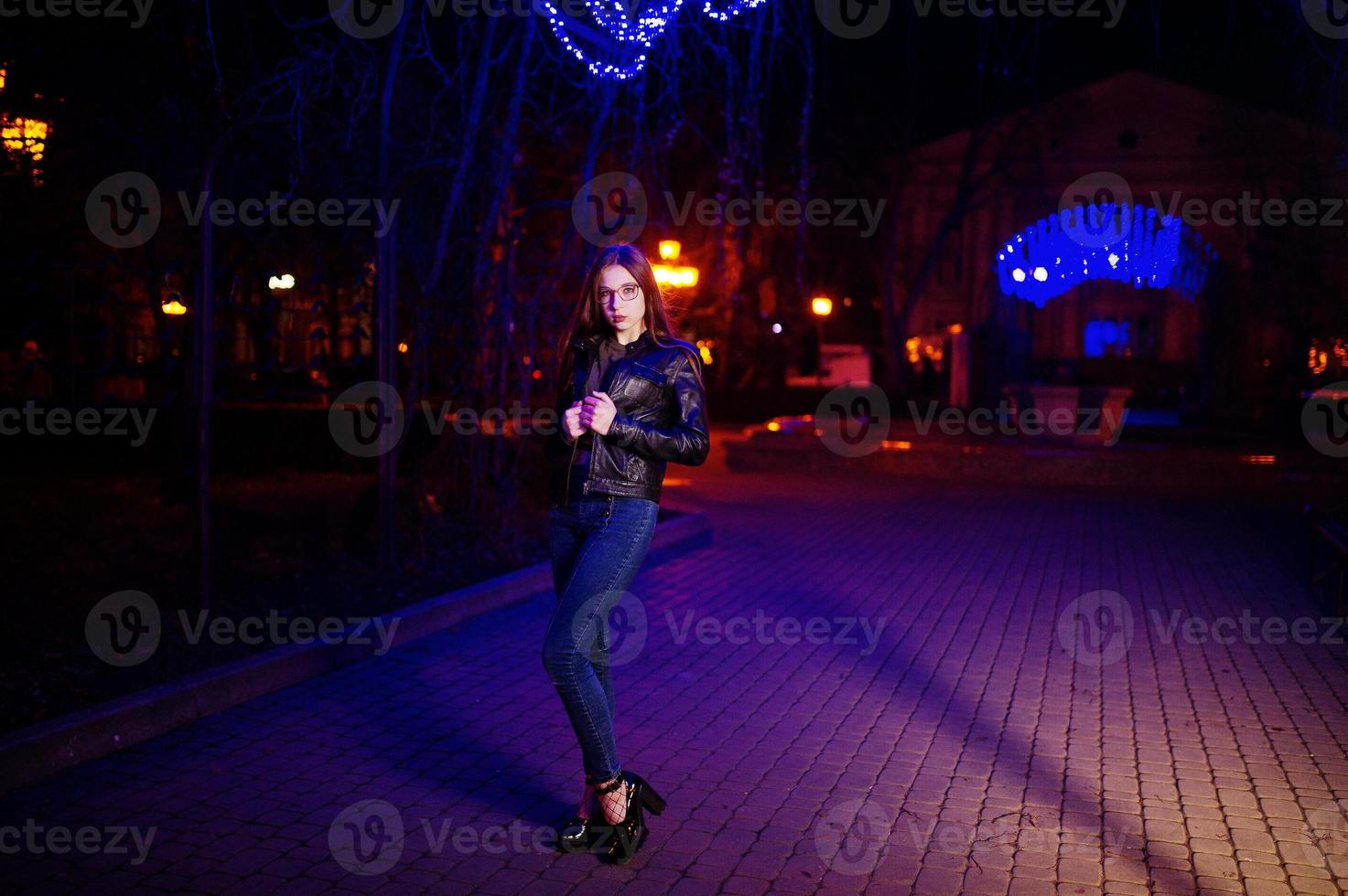 nachtportret van meisjesmodel op bril, jeans en leren jas tegen blauwe lichtenslinger van stadsstraat. foto