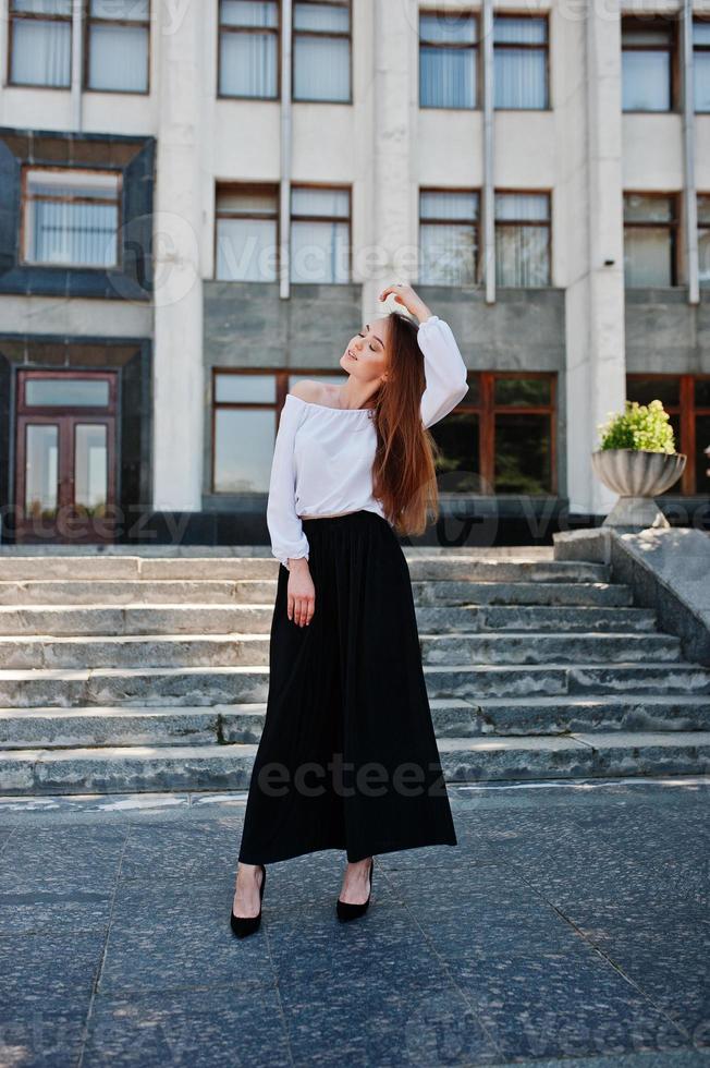 portret van een fantastische jonge succesvolle vrouw in witte blouse en brede zwarte broek poseren op de trap met een enorm wit gebouw op de achtergrond. foto