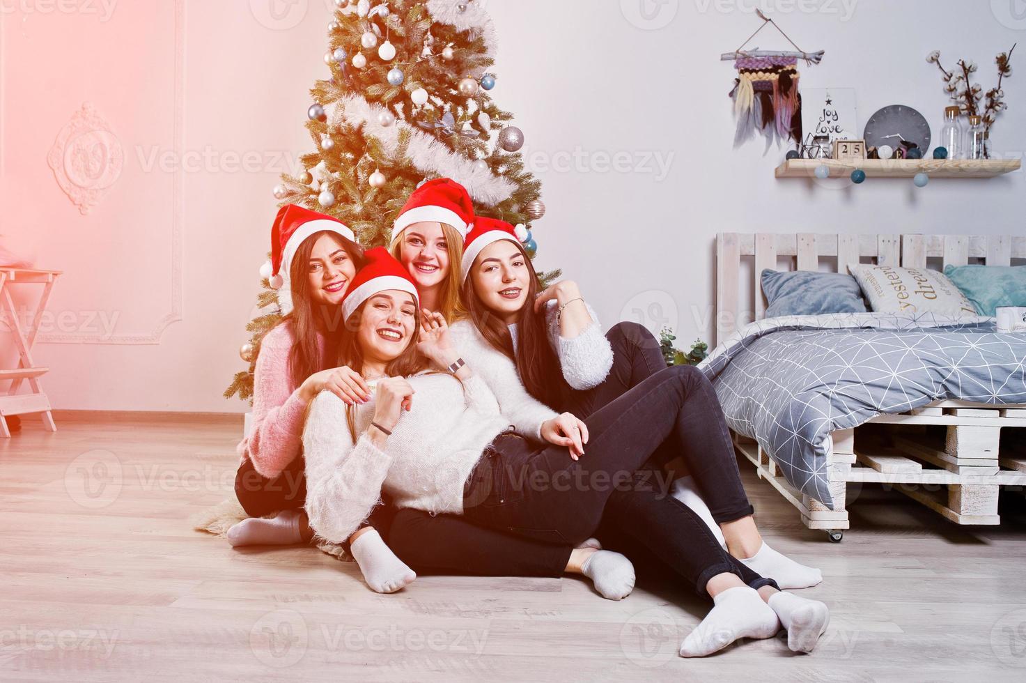 vier schattige vriendenmeisjes dragen warme truien, zwarte broeken en rode kerstmutsen tegen nieuwjaarsboom met kerstversiering in de witte kamer. foto