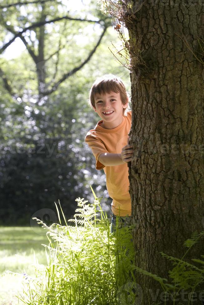 jongen (8-10) gluren achter boom, lachend, portret foto