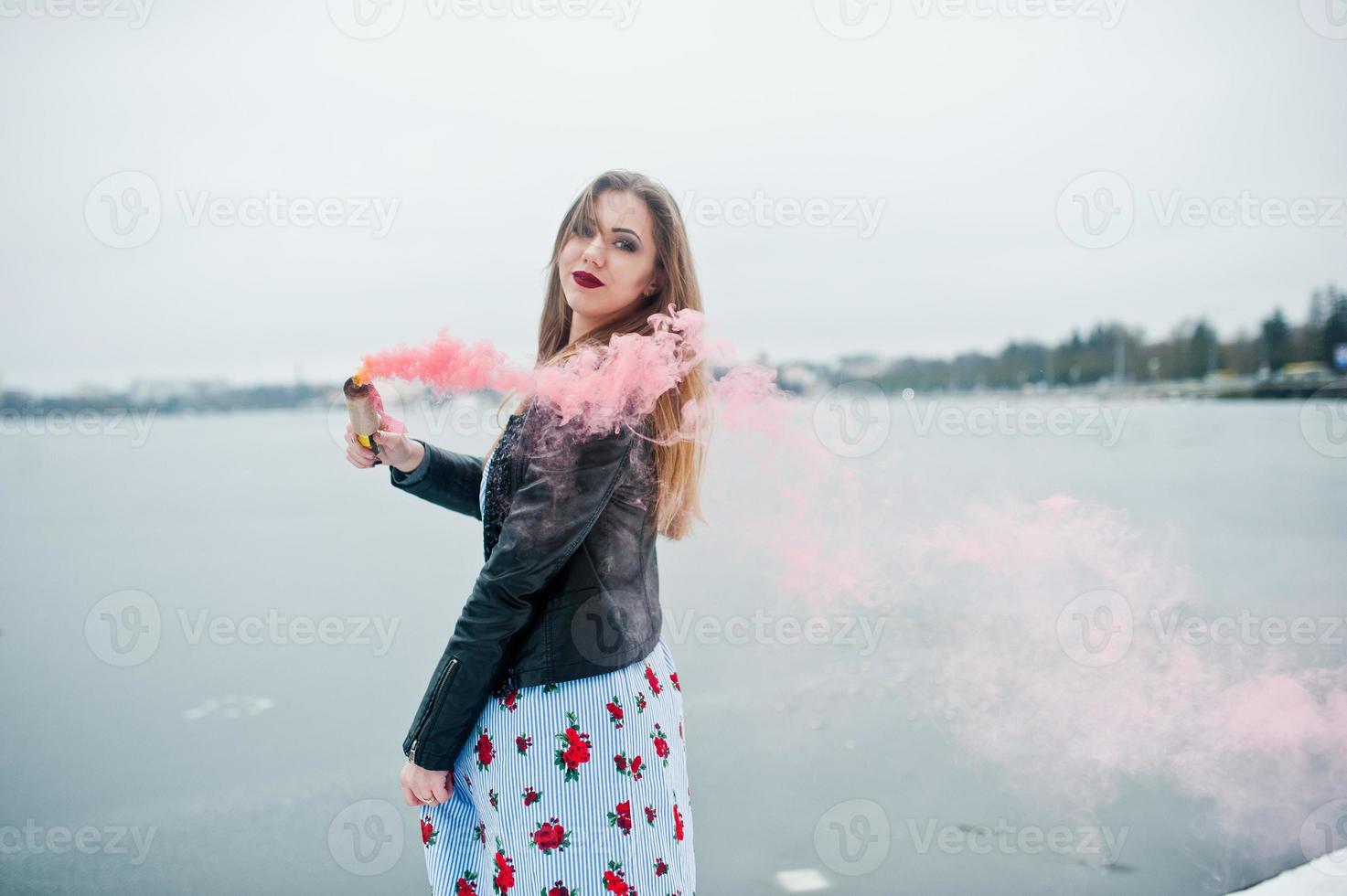 stijlvol meisje in leren jas houdt roze rookflare op winterdag tegen bevroren meer. foto