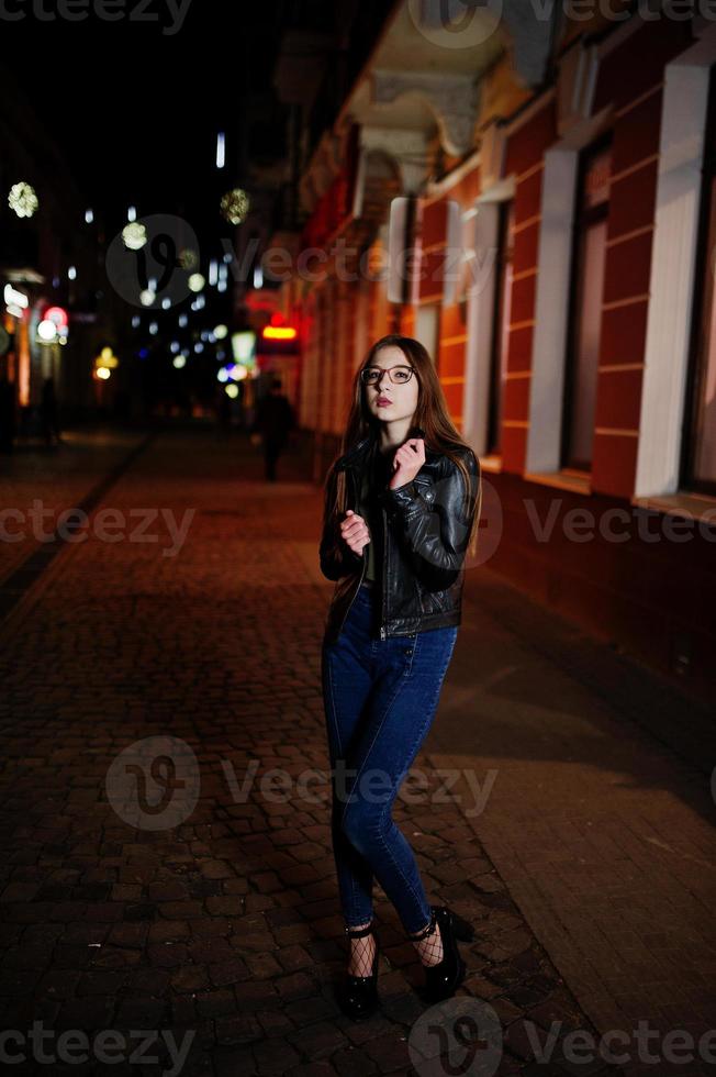 nachtportret van meisjesmodel op bril, jeans en leren jas, met rugzak, tegen stadsstraten. foto