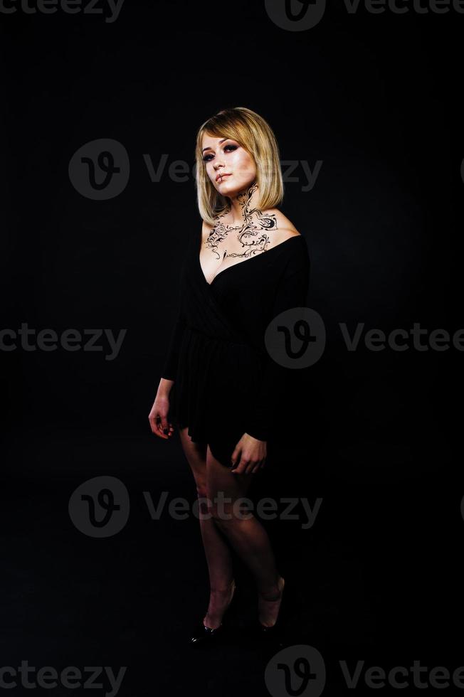 studioportret van blond meisje met oorspronkelijk make-up op nek, draag op zwarte jurk op donkere achtergrond. foto