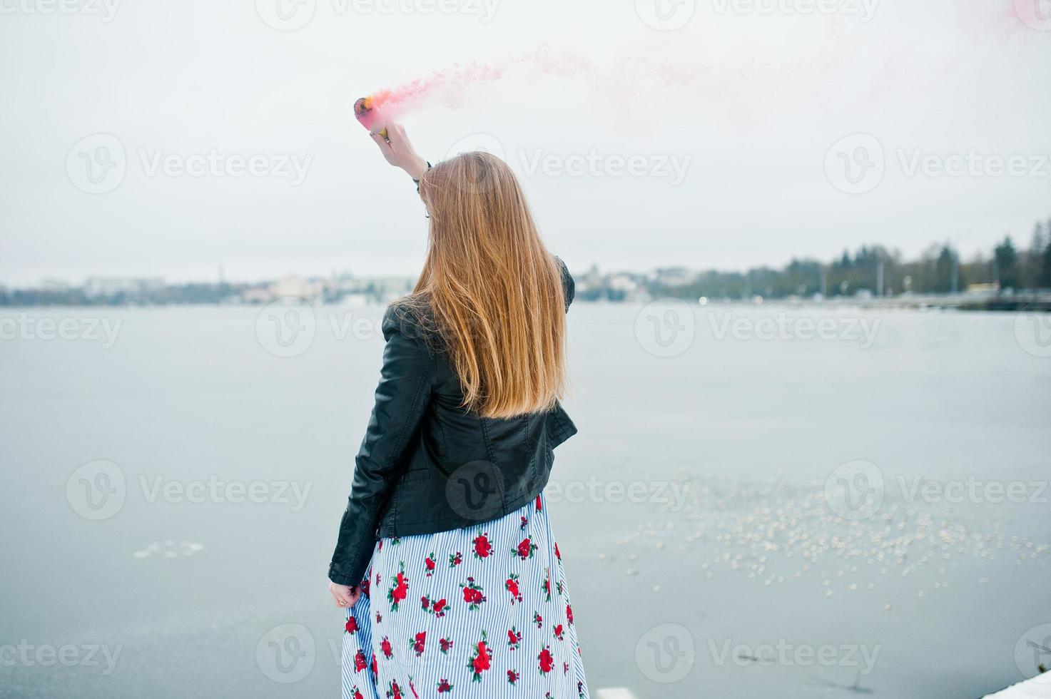 stijlvol meisje in leren jas houdt roze rookflare op winterdag tegen bevroren meer. foto