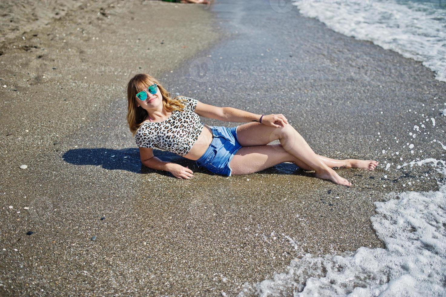 mooi model ontspannen op een strand van zee, het dragen van een korte jeans, luipaard shirt en zonnebril. foto