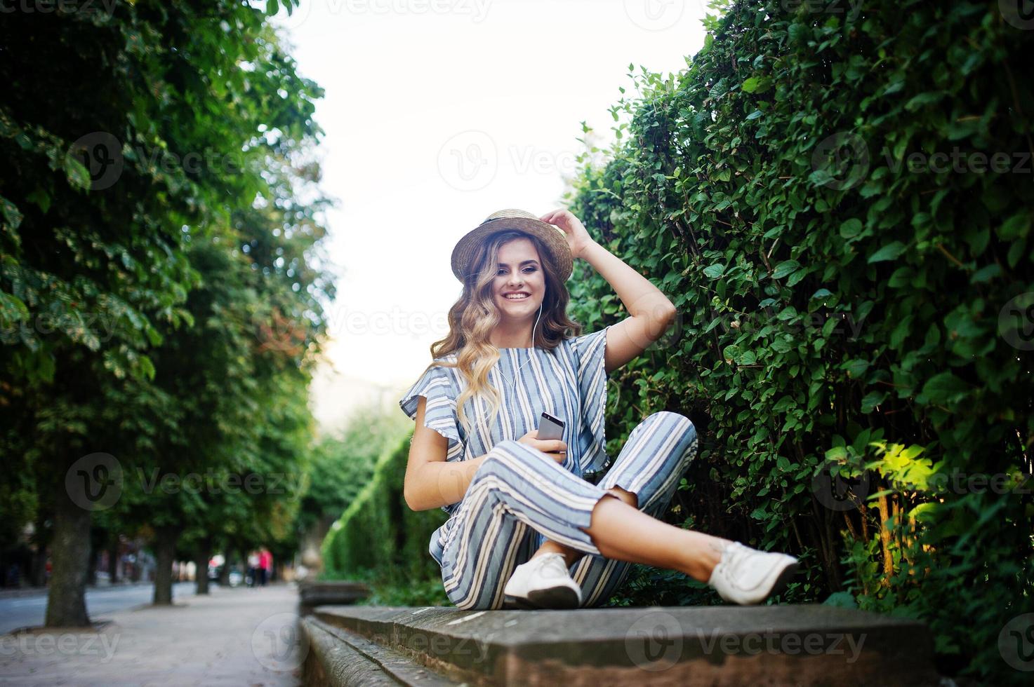 portret van een prachtige jonge vrouw in gestreepte overall zittend in het park en luisterend naar de muziek met haar koptelefoon op. foto