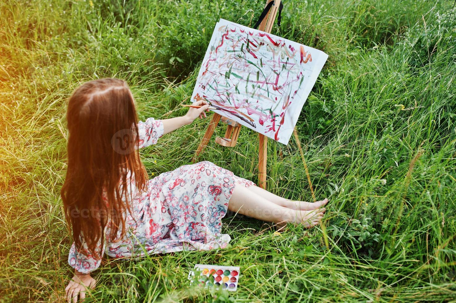 portret van een prachtige gelukkige jonge vrouw in een mooie jurk zittend op het gras en schilderen op papier met aquarellen. foto