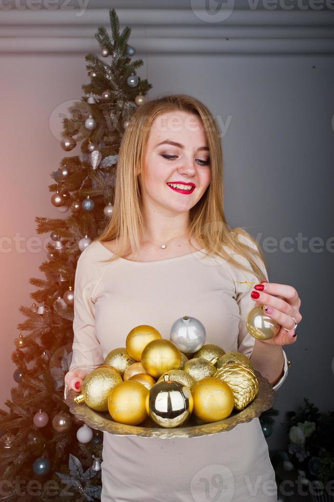 schattig blond meisje op beige jurk tegen nieuwjaarsboom met gouden nieuwjaarsballen in de studio. fijne vakantie. foto