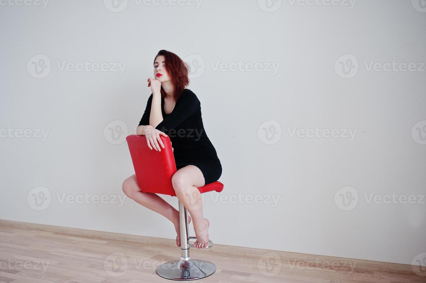roodharige meisje op zwarte jurk tuniek zittend op rode stoel tegen witte muur op lege kamer. foto