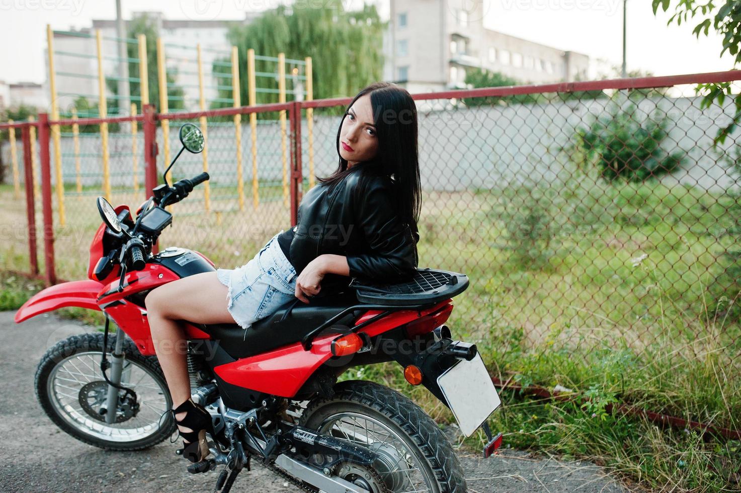 portret van een coole en geweldige vrouw in jurk en zwart leren jack zittend op een coole rode motor. foto