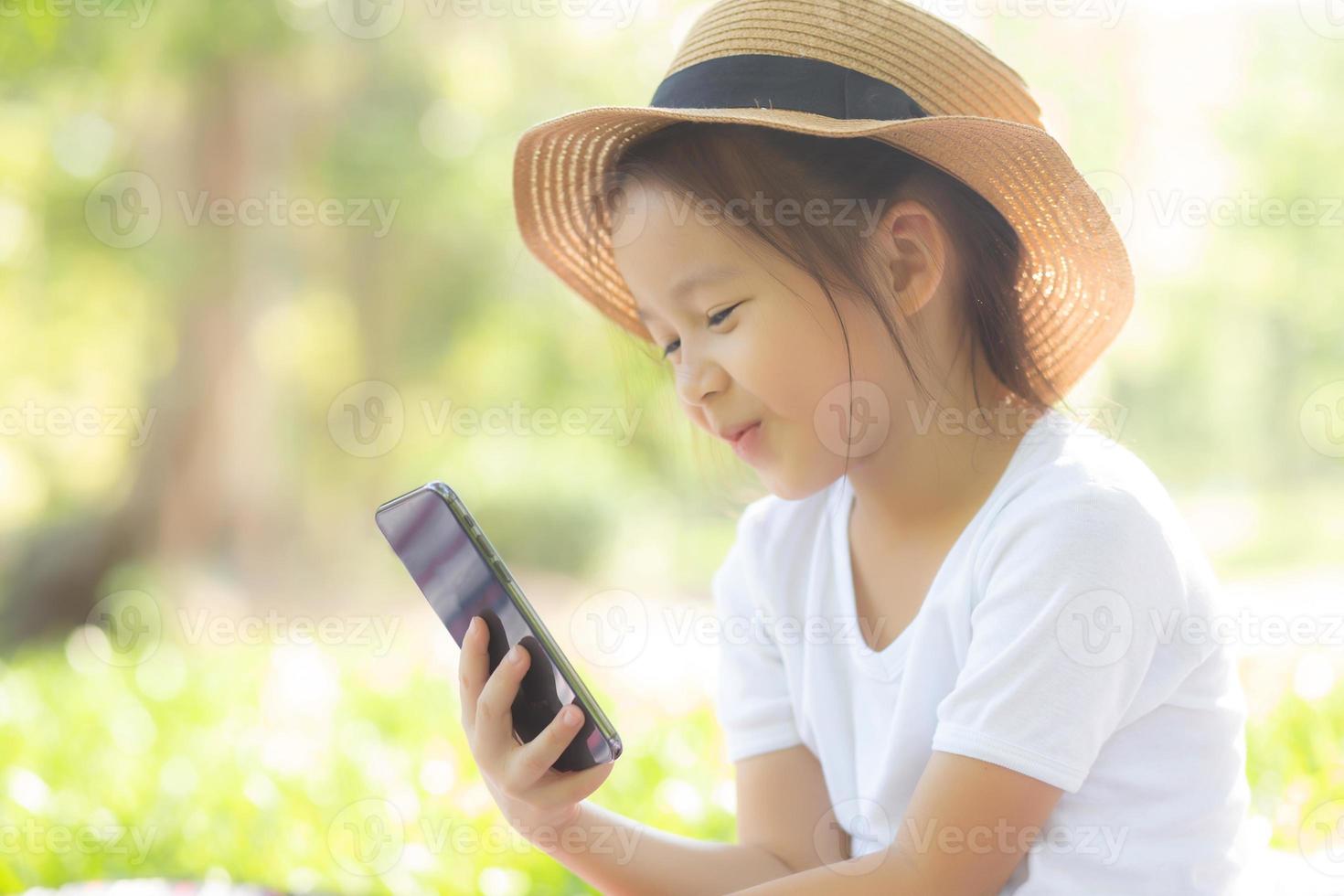 mooi Aziatisch kind glimlachend met behulp van slimme mobiele telefoon in de tuin, kind heeft passie voor het spelen van digitale games op smartphone op internetnetwerk in het park met happy, lifestyle en technologie concept. foto