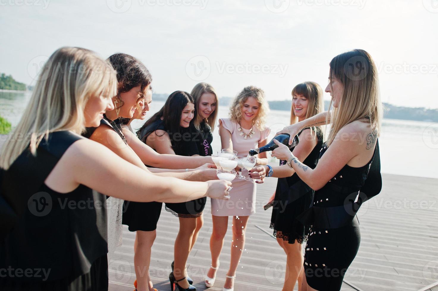 groep van 8 meisjes dragen op zwart en 2 bruiden op vrijgezellenfeest tegen zonnig strand champagne drinken. foto