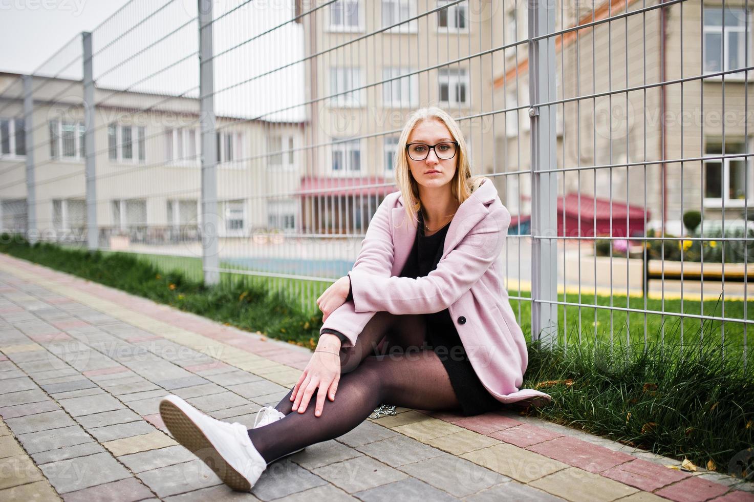 blond meisje met bril en roze jas, zwarte tuniek zittend tegen hek op straat. foto