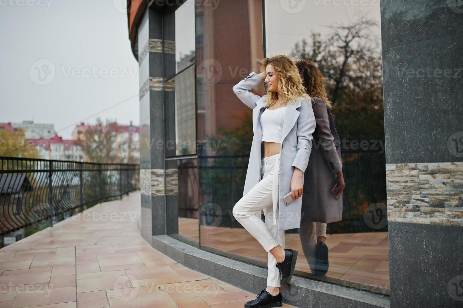 stijlvolle krullend blond model meisje slijtage op wit met mobiele telefoon bij de hand poseren tegen groot raam. foto