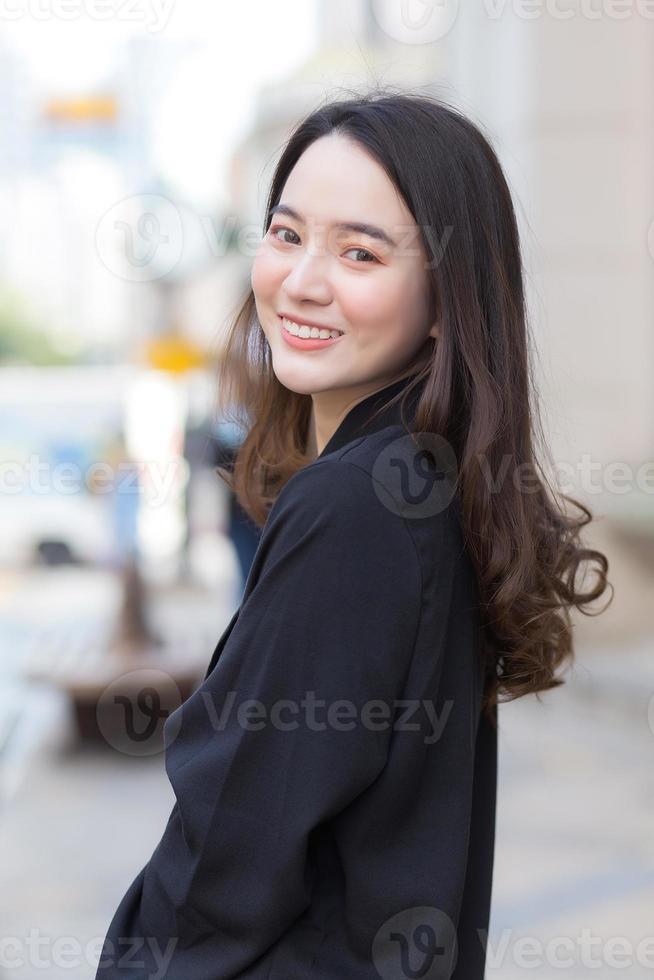 een portret van een mooie, langharige Aziatische vrouw in een zwarte jas die buiten in de stad loopt en glimlacht. foto