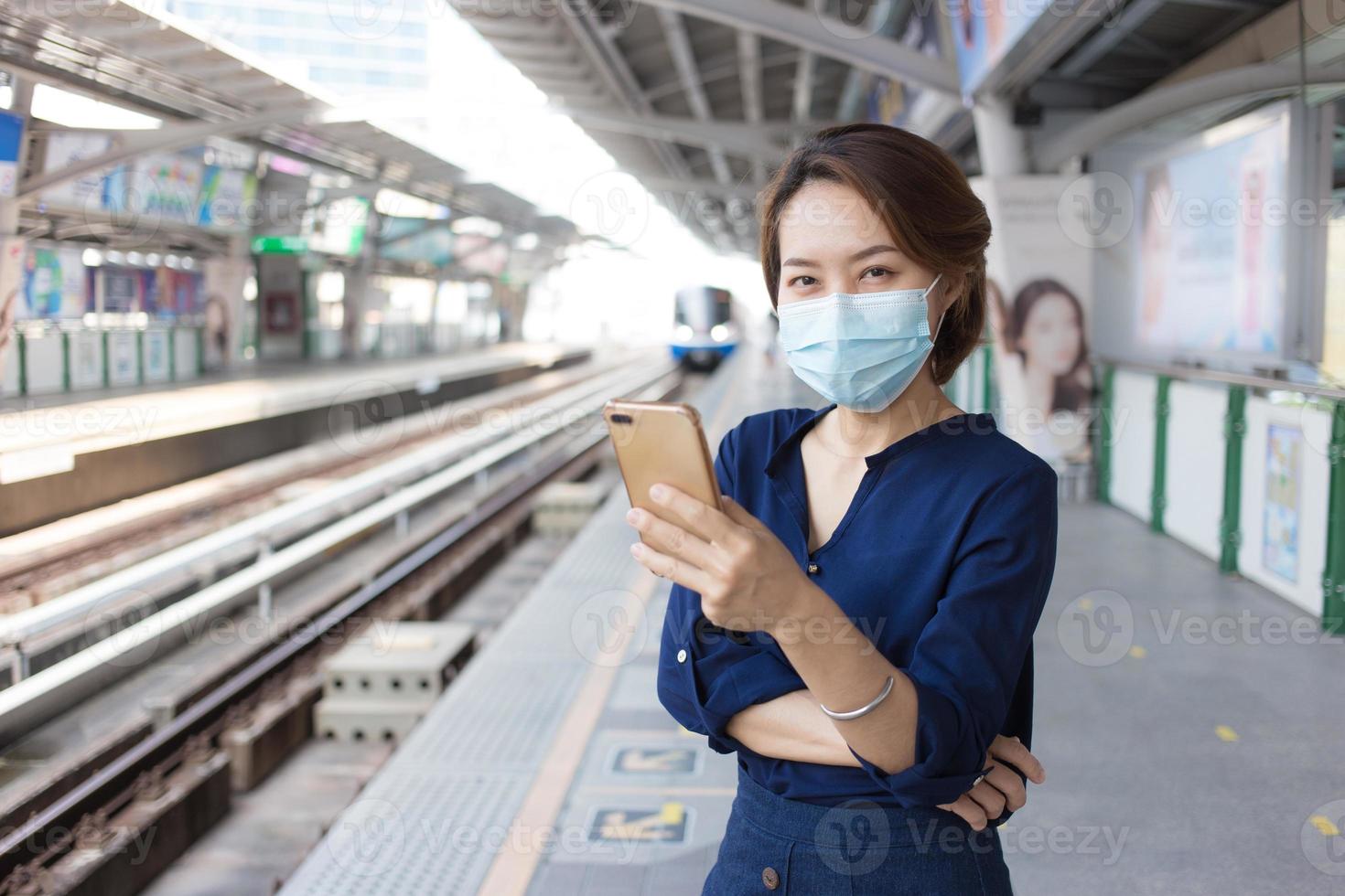 aziatische dame met kort haar die een smartphone speelt tijdens het wachten op de trein op het station om te reizen en een medisch gezichtsmasker draagt als een nieuwe normale levensstijl. foto