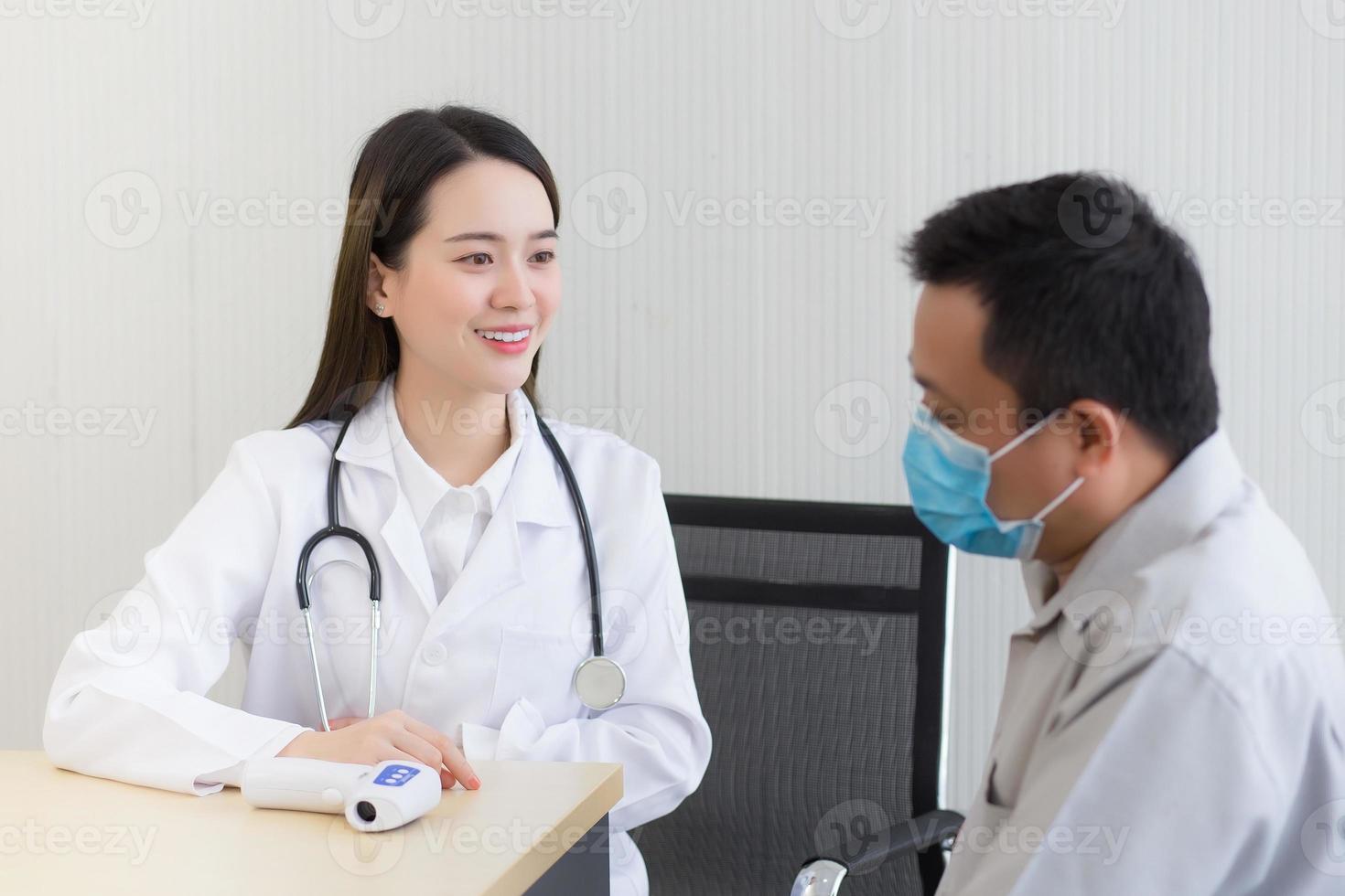 aziatische mooie jonge vrouwelijke arts die met een man-patiënt praat over zijn pijn en symptoom terwijl ze een gezichtsmasker opzetten om coronavirusziekte en thermometer op tafel in het ziekenhuis te voorkomen. foto