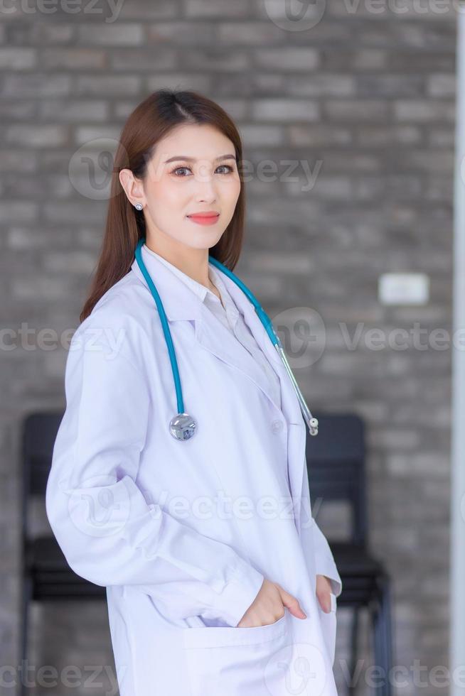 Aziatische vrouw arts permanent met hand in zak blij en glimlach in het ziekenhuis. het dragen van een wit gewaad en een stethoscoop. nieuw normaal en zorgconcept. foto