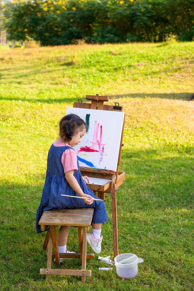 een klein meisje zit op de houten bank en schildert op het canvas dat op een tekenstandaard is geplaatst foto