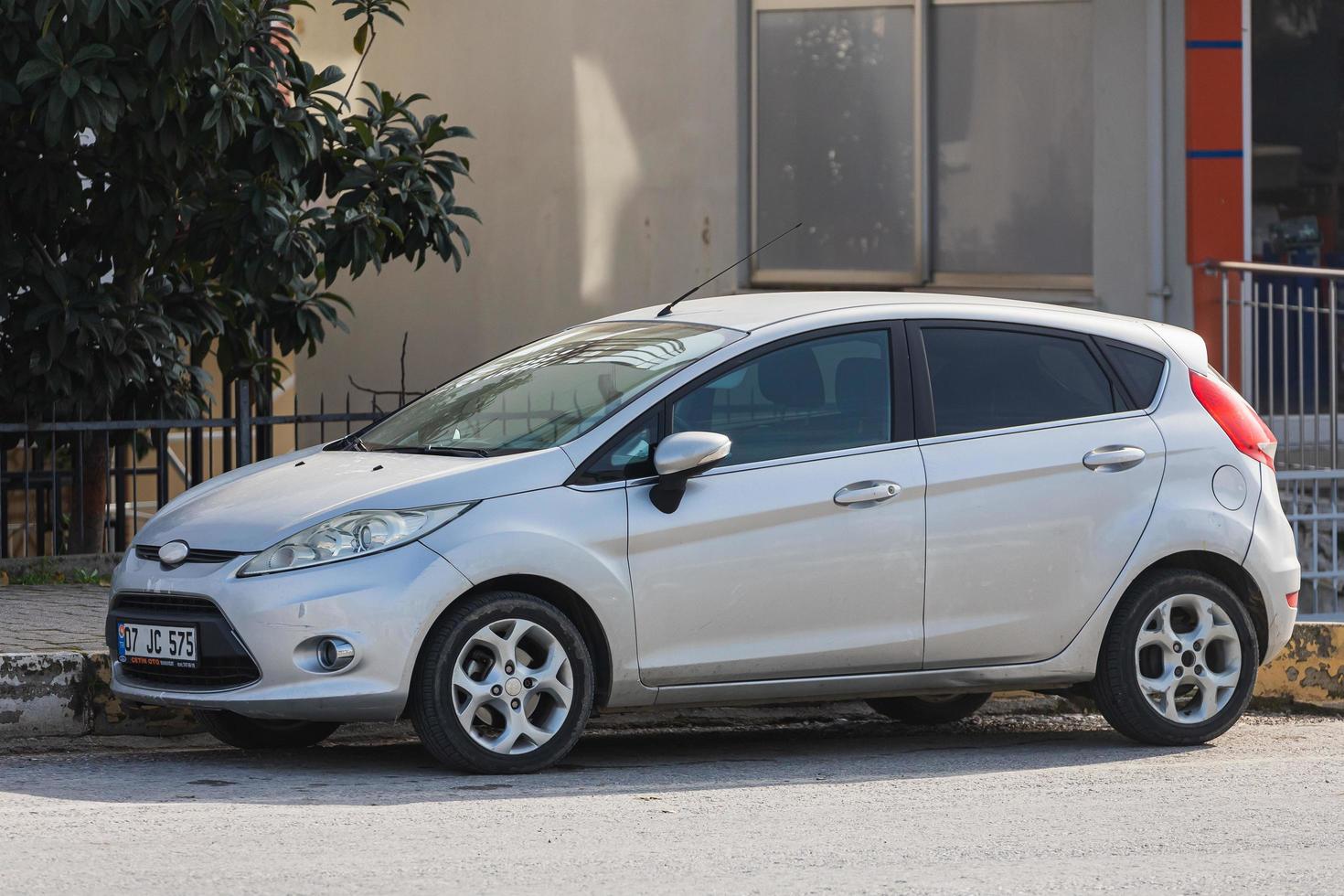 Side Turkije 18 februari 2022 Silver Ford Fiesta wordt op een warme dag op straat geparkeerd tegen de achtergrond van een buildung, park, hek, winkels foto