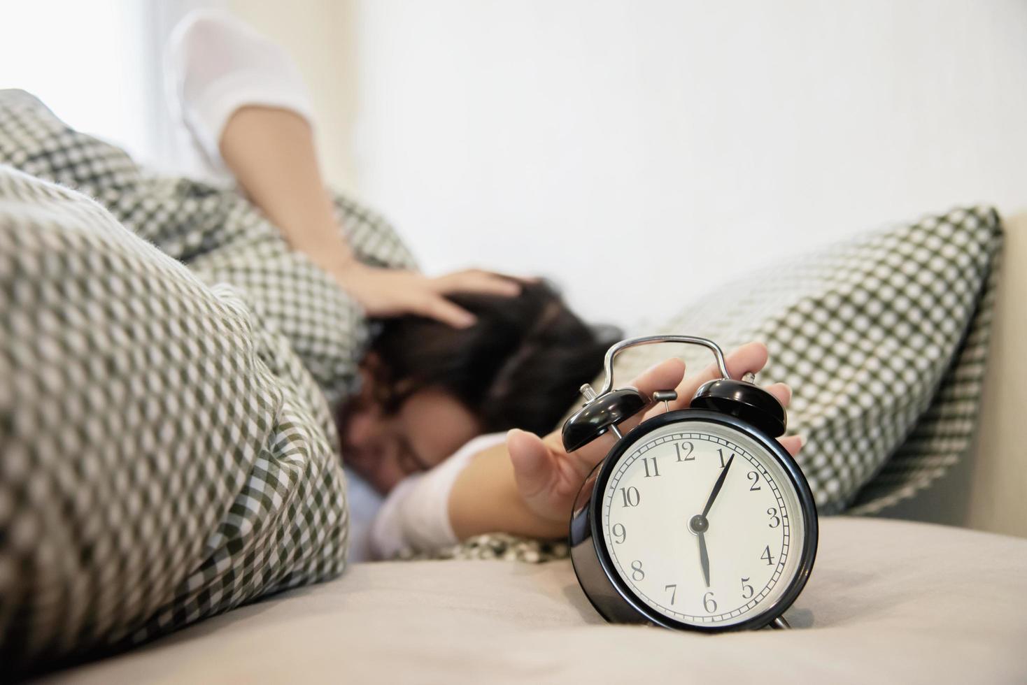 slaperige vrouw die 's ochtends de wekker vasthoudt met laat wakker worden - concept van het dagelijks leven thuis foto