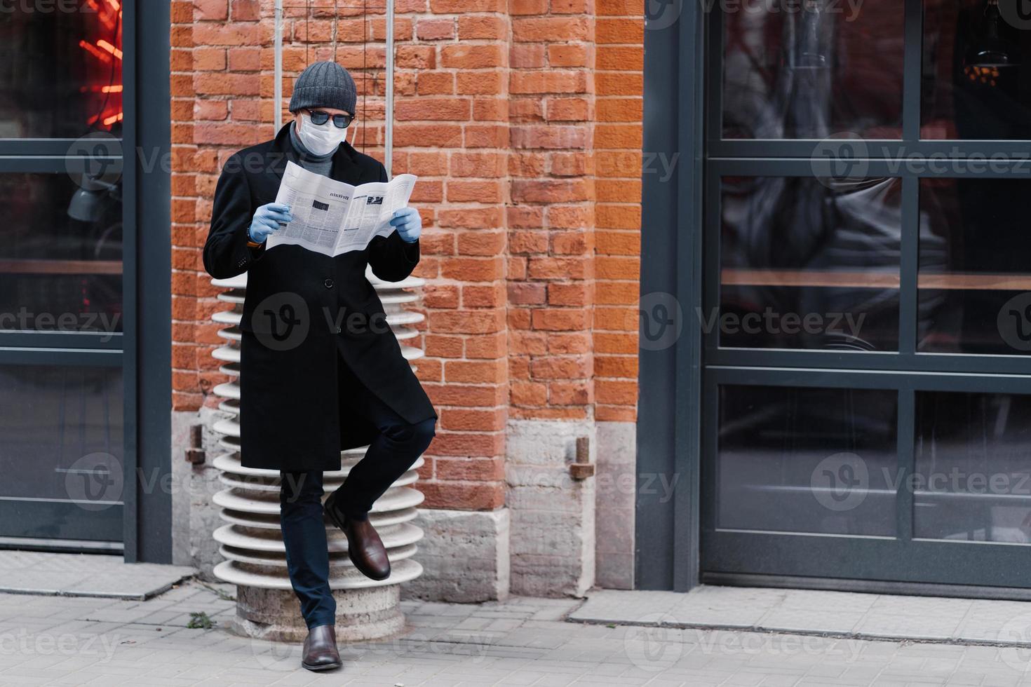 volledige opname van een serieuze man gekleed in een zwarte jas, draagt een zonnebril en een medisch masker, leest de krant, poseert tegen een bakstenen muur, voorkomt dat hij coronavirus krijgt. epidemie en quarantaine foto