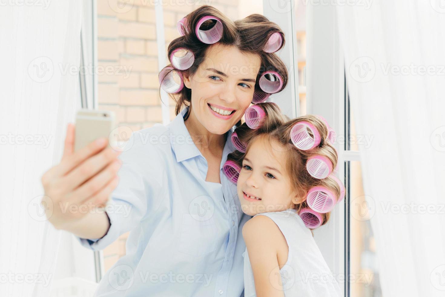 gelukkige vrouwelijke moeder en haar kleine kind met krulspelden op hoofd, poseren voor het maken van selfie, moderne smartphone gebruiken, bij het raam staan, voorbereiden op vakantie. gelukkige familie, technologie, lifestyle concept foto