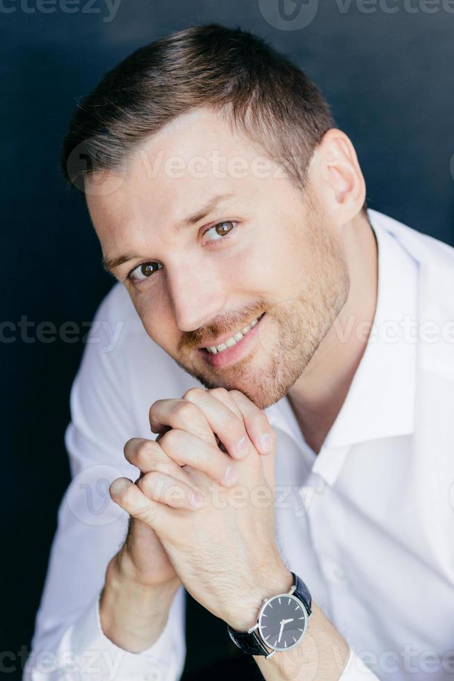 verticaal portret van knappe, vrolijke ongeschoren man met vriendelijke glimlach, houdt de handpalmen tegen elkaar gedrukt onder de kin, draagt een horloge bij de hand, kijkt vrolijk naar de camera. professionele jonge man werknemer foto