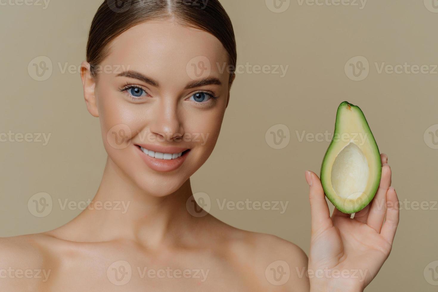 close-up shot van topless jonge blanke vrouwelijke model heeft donker gekamd haar glimlacht zachtjes houdt de helft van avocado staat shirtless indoor geeft om huid en teint. schoonheid wellness-concept foto