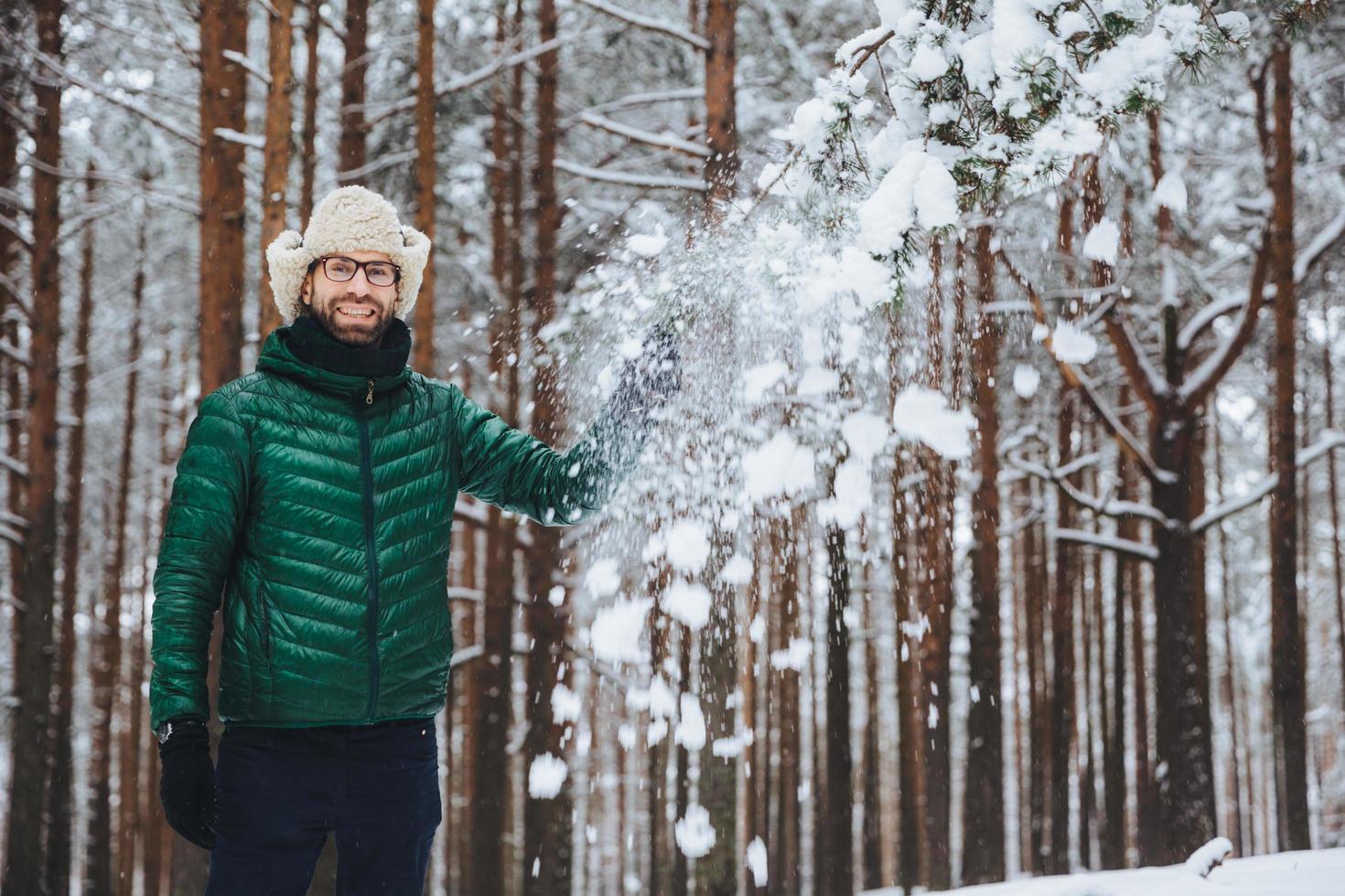 lachende heerlijke man gekleed in warme kleding, staat in het winterbos, gooit sneeuw in de lucht, heeft alleen plezier, heeft een goed humeur, drukt positieve emoties en gevoelens uit. positiviteit concept foto