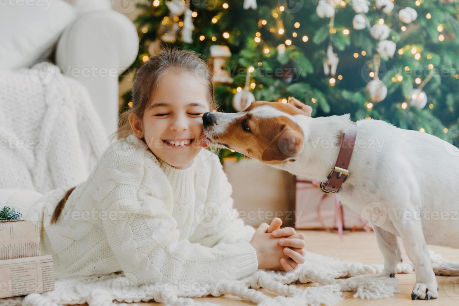 mooie puppy likt chillds gezicht veel plezier samen poseren op de vloer in een gezellige kamer tegen versierde dennenboom, huidige dozen. gelukkig nieuwjaarsconcept. begin van de wintervakantie. kerstvoorbereiding foto