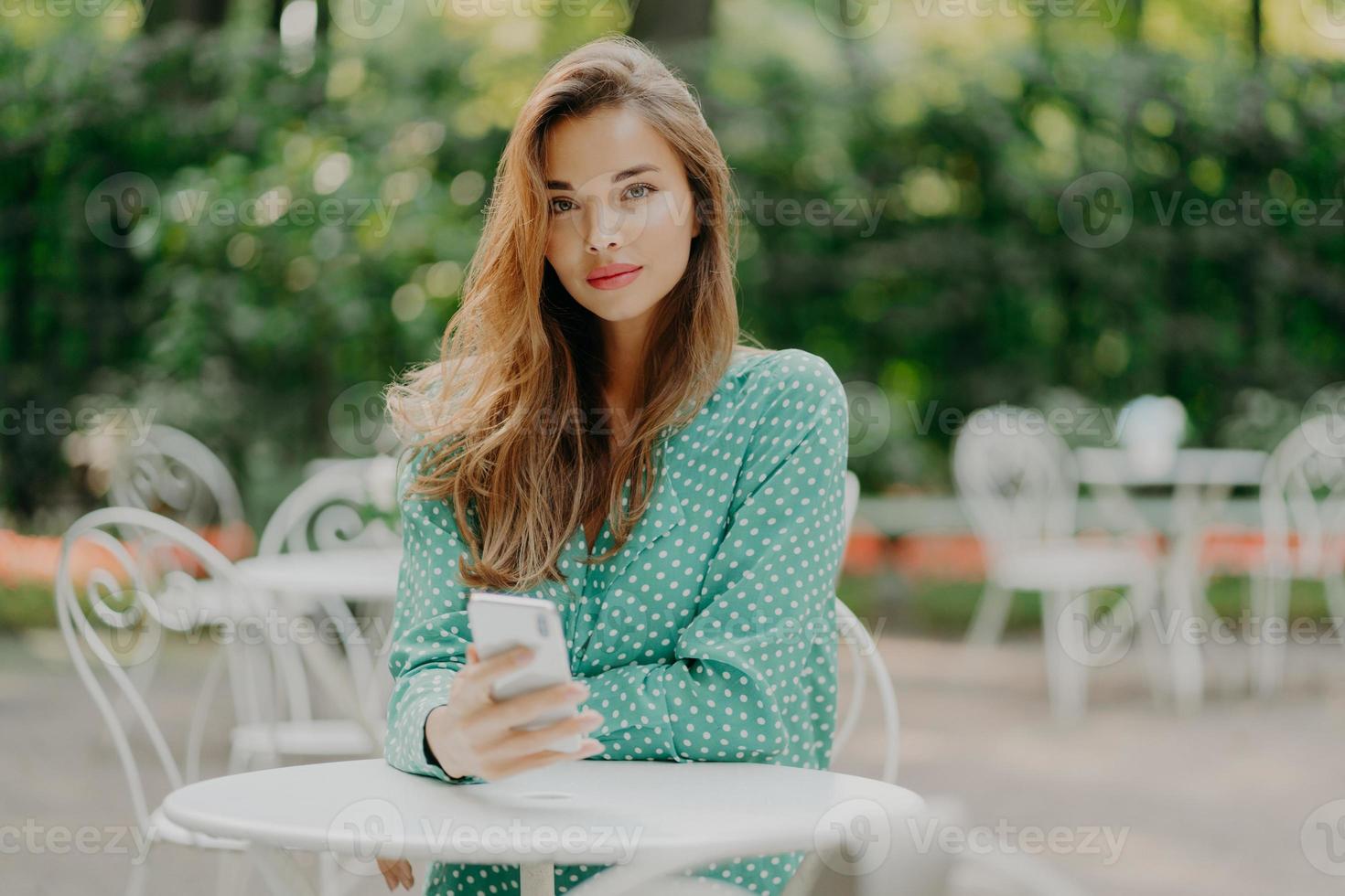 buitenopname van modieuze vrouw met lang haar, gekleed in groen polkadot-shirt, gebruikt moderne mobiele telefoon voor communicatie, verbonden met internet, zit aan witte tafel in buitencafé, wacht op bestelling foto