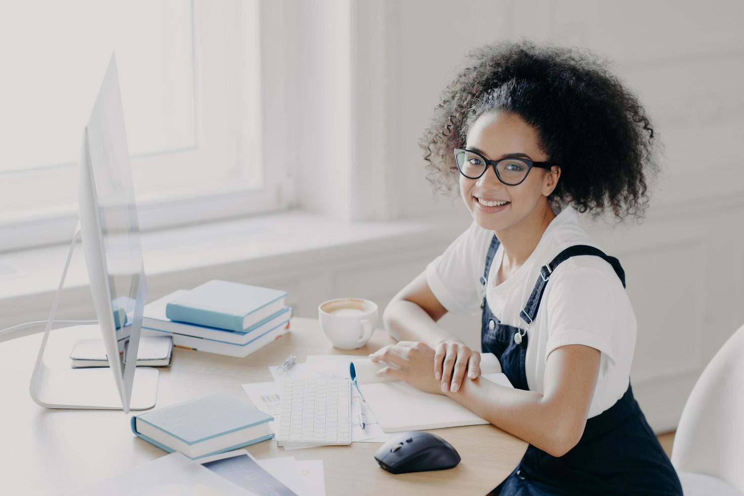 positieve afro-amerikaanse vrouwelijke freelancer poseert op het werk met papieren en schoolboeken, werkt op afstand op de computer, heeft een koffiepauze, werkt in haar eigen kast, draagt vrijetijdskleding. mensen, baan foto