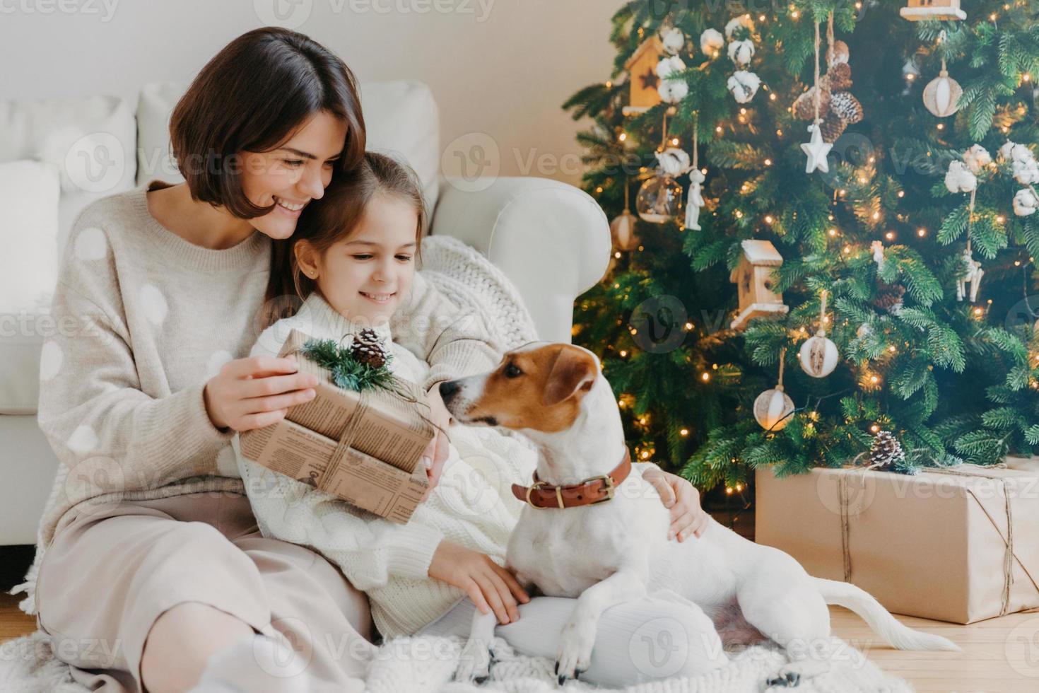 gelukkige jonge vrouw omhelst haar dochtertje, houdt kerstcadeau, wacht op wintervakantie, spelen met stamboom puppy, poseren op de vloer in de buurt van versierde nieuwjaarsboom. gelukkig familie tijd concept foto