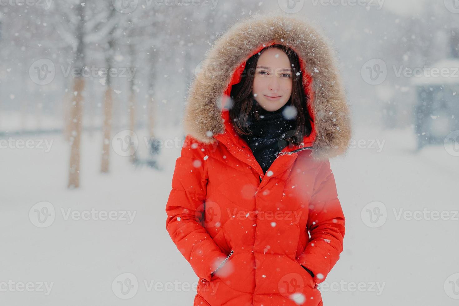 mooi vrouwelijk model in rode jas, stads buiten tijdens sneeuwval, kijkt met donkere ogen naar camera, gaat wandelen met vriendje, ademt frisse lucht. mensen, wintertijd en vrije tijd concept foto