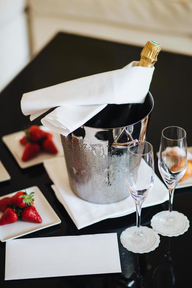 fles champagne in emmer met ijs en lege glazen op zwarte tafel met heerlijke aardbei. geserveerd tafel met dessert en alcoholische drank voor speciale gelegenheid foto