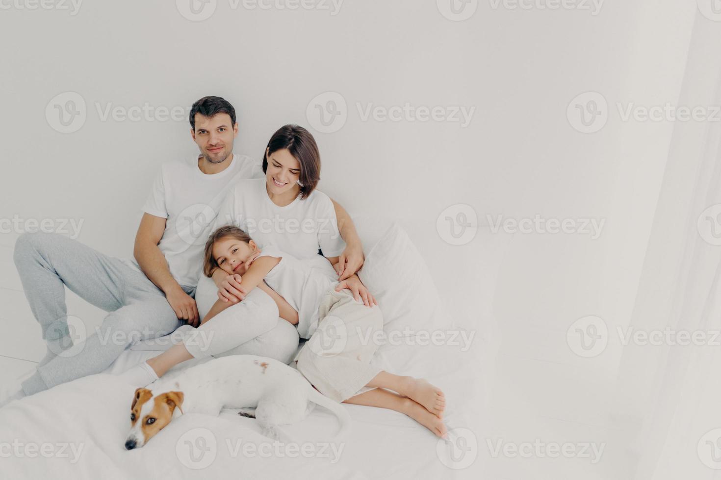 gelukkige familie pose op wit bed tijdens het weekend. vader, moeder, hun dochter en hond voelen zich op hun gemak in de slaapkamer, omhelzen en hebben plezier, ontspannen en vrolijk zijn. vriendelijke familie en rust foto