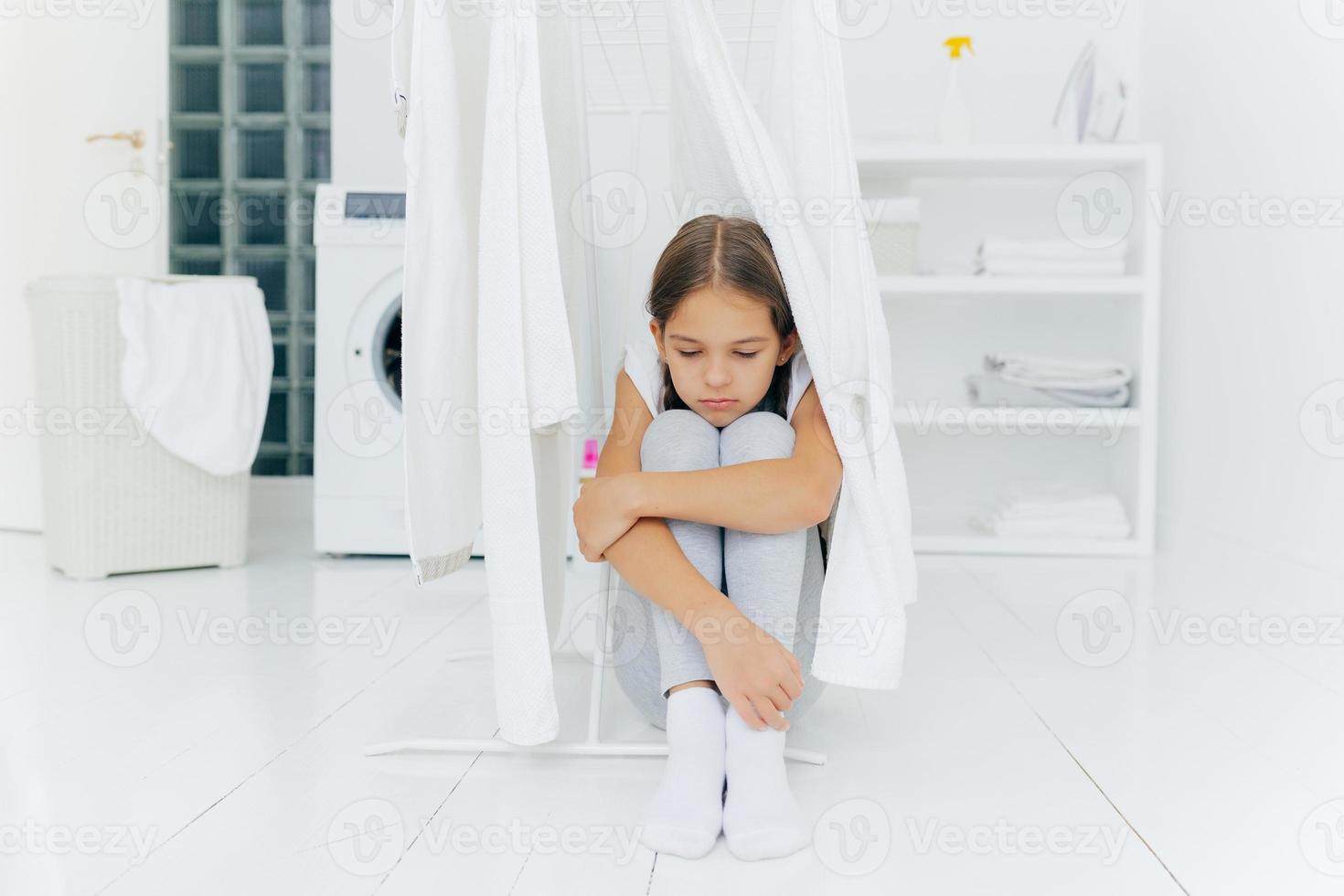 klein schattig meisje zit op de vloer, wordt gestraft door ouders, poseert in de buurt van wasdroger, gefocust met droevige uitdrukking, wasmachine, mand met wasgoed en console, denkt over iets na foto