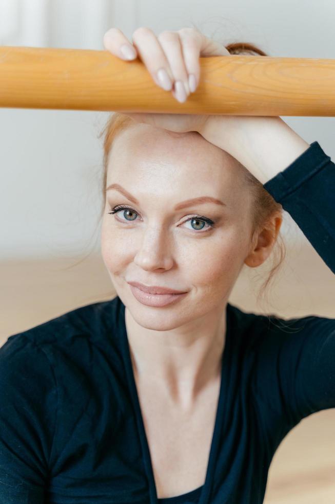 verticaal schot van kalme frisse vrouw met een gezonde huid, gemberhaar, heeft make-up, draagt zwarte kleding, houdt de hand op de barres, kijkt recht naar de camera met blauwe ogen. danseres traint in hal foto