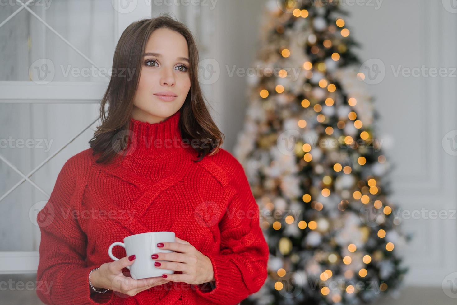 indoor shot van bedachtzame jonge vrouw in warme winterkleren, geniet van warme koffie of cappuccino, kijkt peinzend opzij, staat in de buurt van versierde kerstboom, heeft rode manicure. gezelligheid concept foto