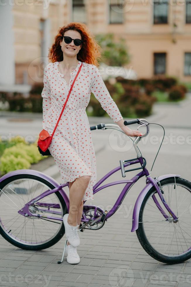 blije vrouw rijdt op de fiets, gekleed in modieuze outfit, glimlacht vrolijk, staat in de buurt van de fiets en wordt in volle lengte gefotografeerd tegen een wazige stadsachtergrond. recreatie en hobby concept foto