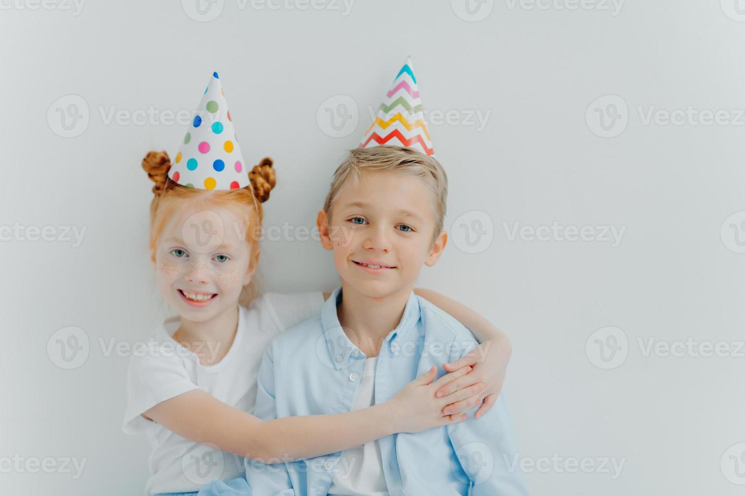 lief roodharig meisje omhelst met liefde haar oudere broer, feliciteert met verjaardag, draagt kegelvormige feestmutsen, heeft een goed humeur op feest, geïsoleerd op witte achtergrond, heeft een vriendschappelijke relatie foto