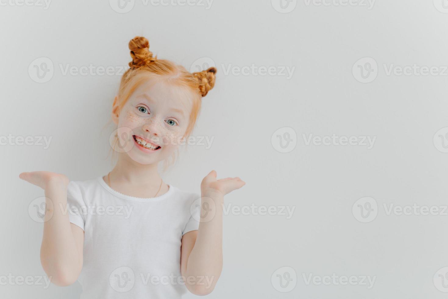 speels roodharig meisje heft handpalmen op, heeft dubbele broodjes, kijkt vrolijk naar de camera, draagt een casual wit t-shirt, poseert binnen, trekt een grappig gezicht, heeft een brede glimlach, een sproetenhuid. kinderen, emoties concept foto