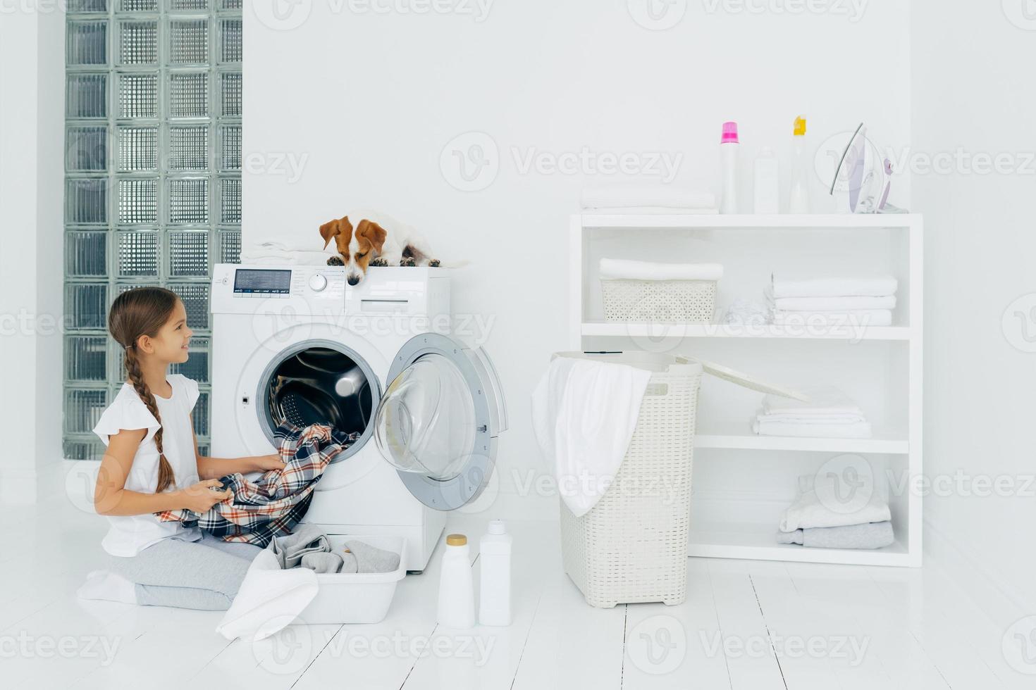 huishouden, kinderen en huishoudelijke taken concept. gelukkig kind laadt de wasmachine uit, legt schone gewassen kleding in de wasbak, nieuwsgierige hond kijkt van bovenaf, ligt op de wasmachine in de wasruimte thuis foto