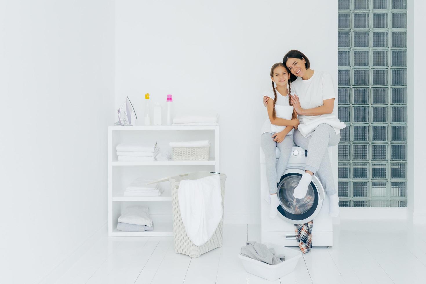 shot van mooie vrouw en zijn kleine dochter omhelzen en glimlachen aangenaam, zitten op de wasmachine, wassen linnen in de wasruimte, hebben een vriendschappelijke relatie, doen thuis de was. huishoudelijk werk concept foto