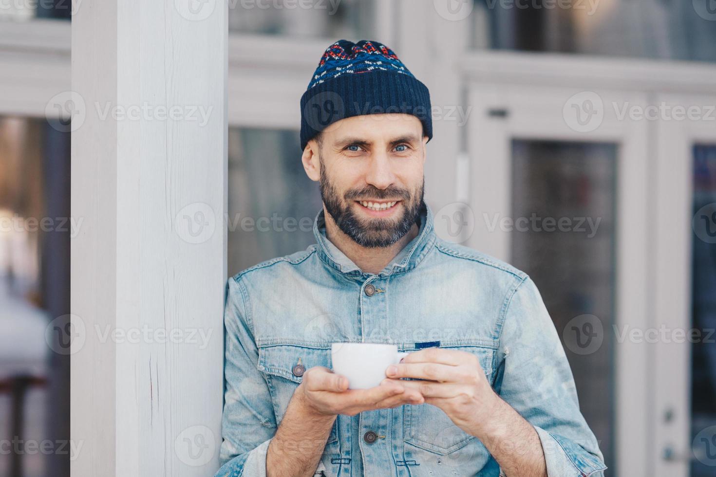 portret van aantrekkelijke bebaarde man heeft een aangename glimlach, gekleed in een spijkerjasje, houdt een witte kop koffie of thee vast, geniet van een goede tijd, heeft een koffiepauze. gelukkige knappe stijlvolle jonge hipster-man foto