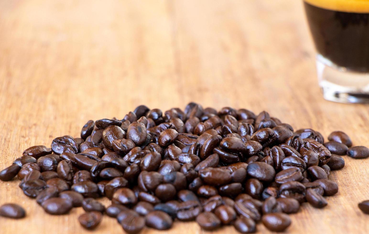 foto van een stapel donkerbruin geroosterde biologische koffieboon op een houten tafel met een zachte focus op een kopje koffie. selectieve focus in het midden van de koffiebonenhoop.