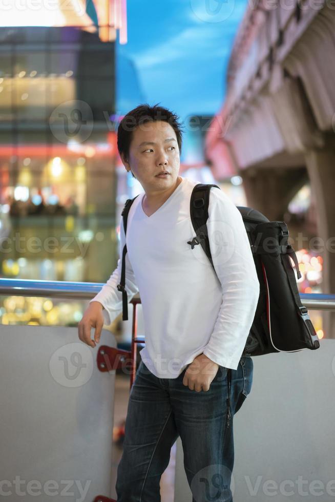 jonge aziatische thaise mannelijke fotograaf met zijn camera en statieftas op zijn rug, plaatst in de schemering in de buurt van bangkok skytrain. foto